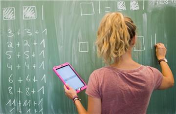  Eine Lehrerin schreibt an eine Schultafel im Mathematikunterricht einer 8. Klasse und blickt dabei auf einen Tablet-Computer iPad.