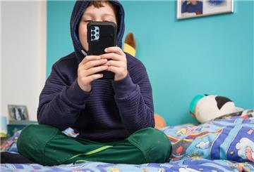  Ein Junge sitzt auf seinem Bett und schaut auf sein Handy.  Trickbetrüger geben sich als Freunde aus – auch auf Kinderhandys.