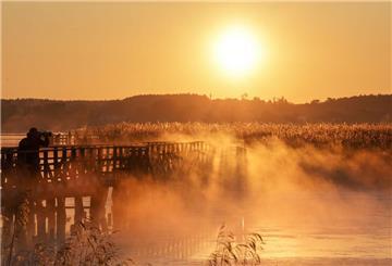 Ein Mann fotografiert kurz nach Sonnenaufgang auf dem 1,5 Kilometer langen Federseesteg den Federsee. Die Sonne färbt den Morgennebel gelblich ein.