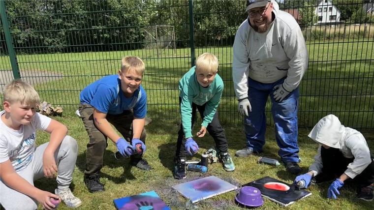 Kinder und ein Betreuer gestalten im Freien kreative Bilder mit Farbspraydosen; sie arbeiten auf dem Rasen vor einem Zaun, umgeben von buntem Material.
