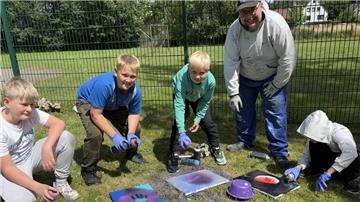 Kinder und ein Betreuer gestalten im Freien kreative Bilder mit Farbspraydosen; sie arbeiten auf dem Rasen vor einem Zaun, umgeben von buntem Material.
