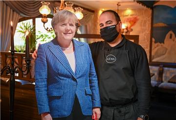 Als Pappaufsteller hat Restaurantbesitzer Alexis Vaiou schon eine Angela Merkel, bei der Lieferung der Wachsfigur der Kanzlerin gibt es unerwartete Komplikationen.