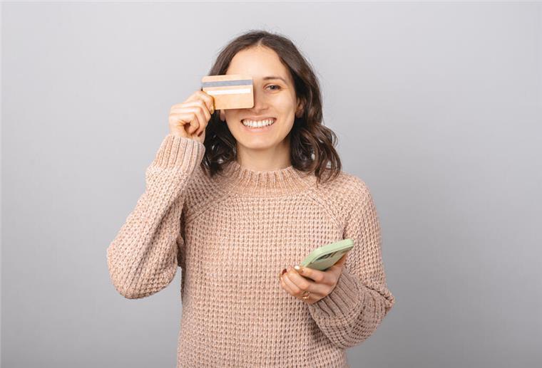 Eine junge Frau hält sich lächelnd ihre Bankkarte vor das Gesicht und ein Smartphone in der Hand.