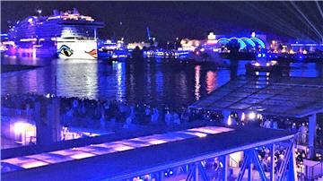 Nachtaufnahme eines Hafens mit einem großen, beleuchteten Kreuzfahrtschiff im Hintergrund, das das charakteristische Design von Augen und Lippen auf der Schiffswand trägt. Der Hafen ist in blauem Licht illuminiert, mit zahlreichen Menschen, die das Szenario von einer Aussichtsplattform und einer unteren Ebene beobachten.