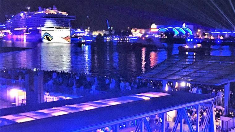 Nachtaufnahme eines Hafens mit einem großen, beleuchteten Kreuzfahrtschiff im Hintergrund, das das charakteristische Design von Augen und Lippen auf der Schiffswand trägt. Der Hafen ist in blauem Licht illuminiert, mit zahlreichen Menschen, die das Szenario von einer Aussichtsplattform und einer unteren Ebene beobachten.
