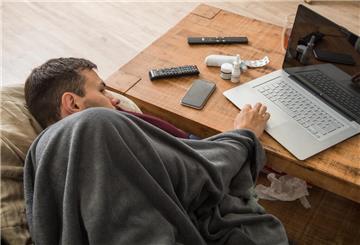 Ein Mann liegt angeschlagen und zugedeckt an einem Tisch, mit einer Hand bedient er einen Laptop. Bei Gesundheitsfragen kann KI helfen – doch fehlerhafte Antworten sind keine Seltenheit.