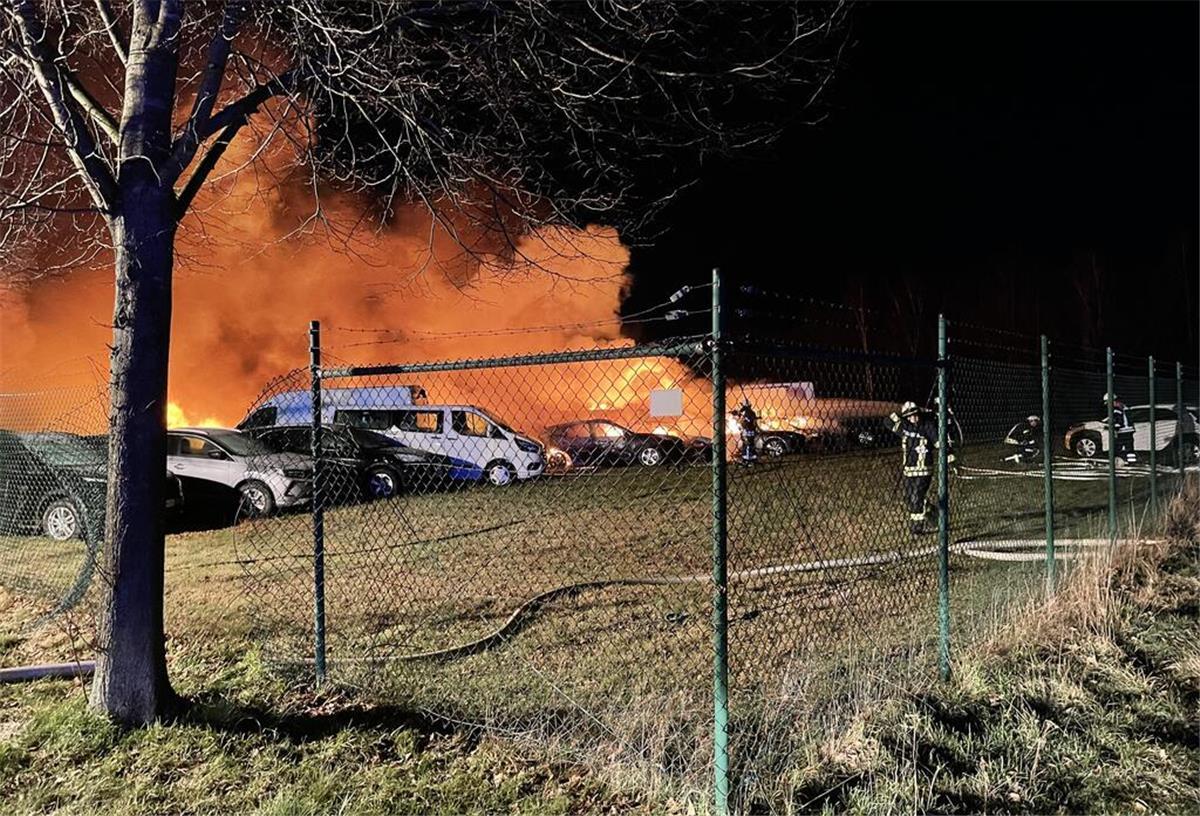 Auf einer eingezäunten Rasenfläche stehen brennende Fahrzeuge kurz nach Eintreffen der Feuerwehr.