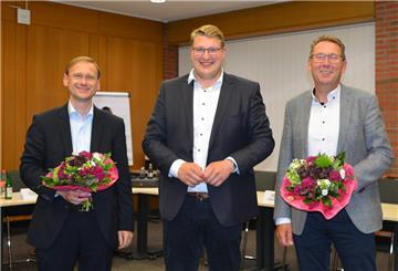 Drei Männer stehen in einem Raum, zwei mit Blumen in der Hand.
