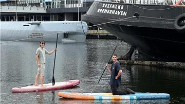 Zwei Männer demonstrieren Stand-up-Paddling in einem Hafen. Im Hintergrund sind ein U-Boot und ein Schiff zu sehen.