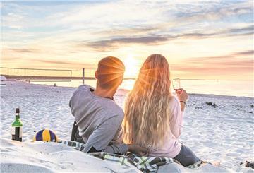Ein Mann und eine Frau liegen angezogen an einem weißen Sandstrand, schauen in die untergehende Sonne und trinken Wein