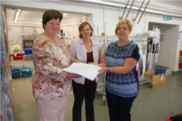 Das verloren gegangene Erbstück ist wieder da: Meike Koopmann (links) und Janine Dieckmeyer übergeben Margarete Hilgen (rechts) in der CVJM-Wäscherei in Nordenham die Tischdecke.