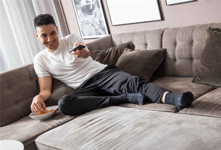 Ein Mann sitzt mit Snacks und einer Fernbedinung in der Hand auf der Couch.