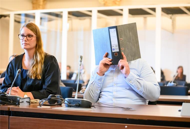 Ein Mann hält in einem Gerichtssaal einen Aktenordner vor das Gesicht. Daneben sitzt eine Frau.