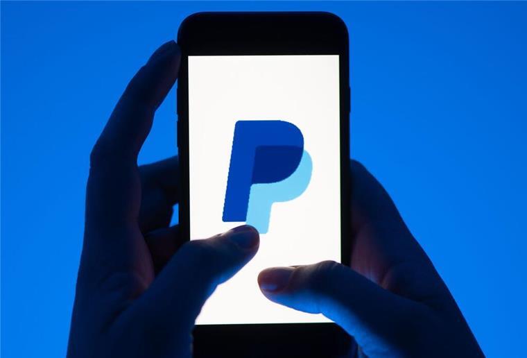 Eine Frau hält ein Smartphone mit dem Logo des Bezahldienstes PayPal. Der Bezahldienst PayPal führt ein Cashback-Programm ein. Doch Kunden sollten sich die AGBs durchlesen, sonst drohen Gebühren.