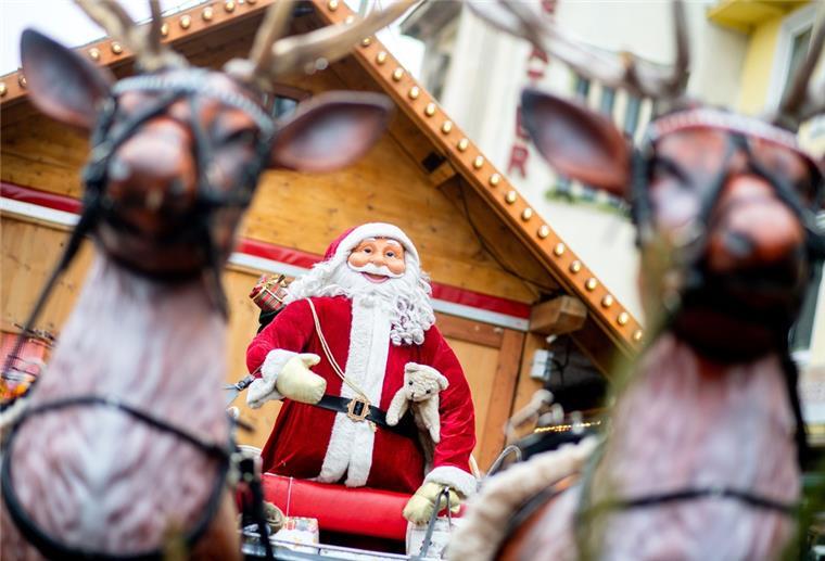 Ein Weihnachtsmann auf einem Schlitten und zwei Rentiere stehen als Dekoration an einem Marktstand auf einem Weihnachtsmarkt.