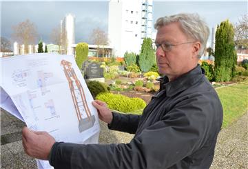 Der Elsdorfer Pastor Michael Grimm steht mit der Zeichnung des geplanten Glockenturms auf dem Friedhof. Der wird voraussichtlich Anfang kommenden Jahres gebaut. Im Hintergrund sind Gebäude der Molkerei zu sehen.