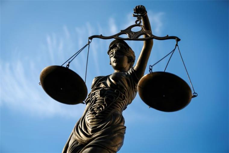 Eine Statue der Justizia mit verbundenen Augen und einer Waage in der Hand steht vor einem blauen Himmel.