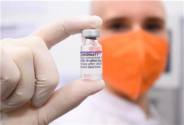 Ein Biontech Impfstoff-Fläschchen wird von jemandem mit orangener Maske in die Kamera gehalten.