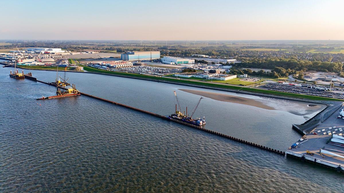Neue Liegeplätze in Cuxhaven: So läuft der Mega-Ausbau am Hafen Luftaufnahme vom Hafen in Cuxhaven mit mehreren Bauschiffen und Großgeräten, die an einer langen Spundwand im Wasser arbeiten. Im Hintergrund sind Lagerhallen, Industrieanlagen und geparkte Fahrzeuge zu sehen.
