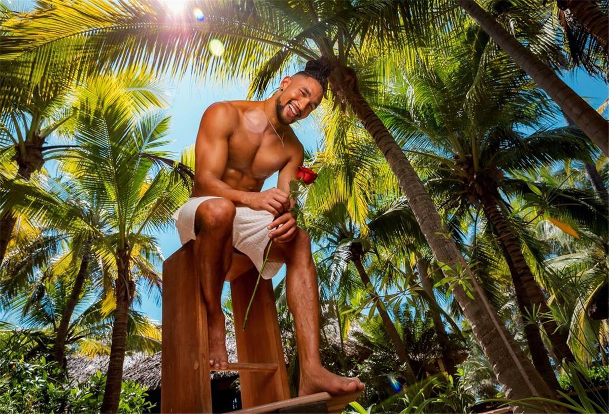 Neuer RTL-"Bachelor" David: Noch nie eine Dating-App genutzt Ein dunkelhäutiger Mann mit Dutt und Rose sitzt unter Palmen und lächelt in die Kamera.