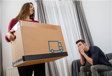 Eine Frau packt ihre Sachen während ihr Mann auf dem Sofa sitzt.