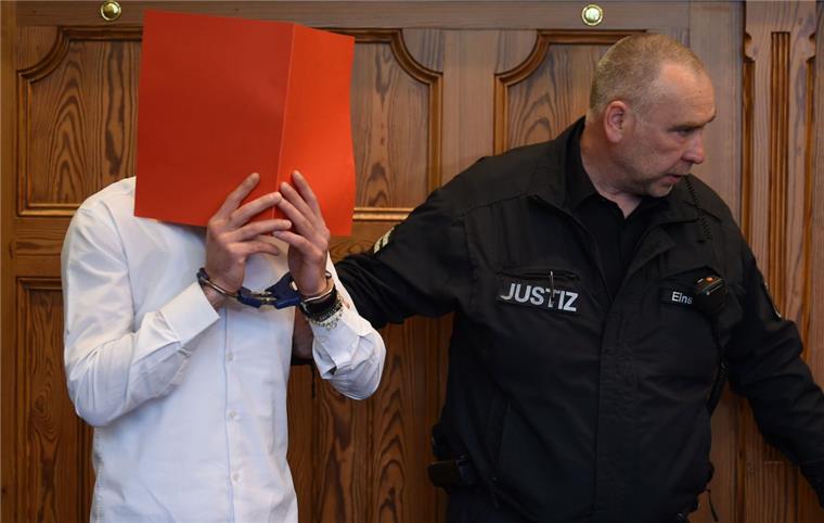Nach Auto-Attacke in Cuxhaven: Vier Jahre Haft für Syrer (29) Der wegen versuchten Mordes Angeklagte wird von einem Justizbeamten im Gerichtssaal auf seinen Platz begleitet und hält sich einen Aktenordner vor das Gesicht.