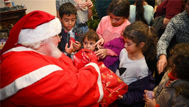 Ein Mann in Weihnachtsmann-Kostüm sitzt vor einer Schar Kinder und überreicht Geschenke.