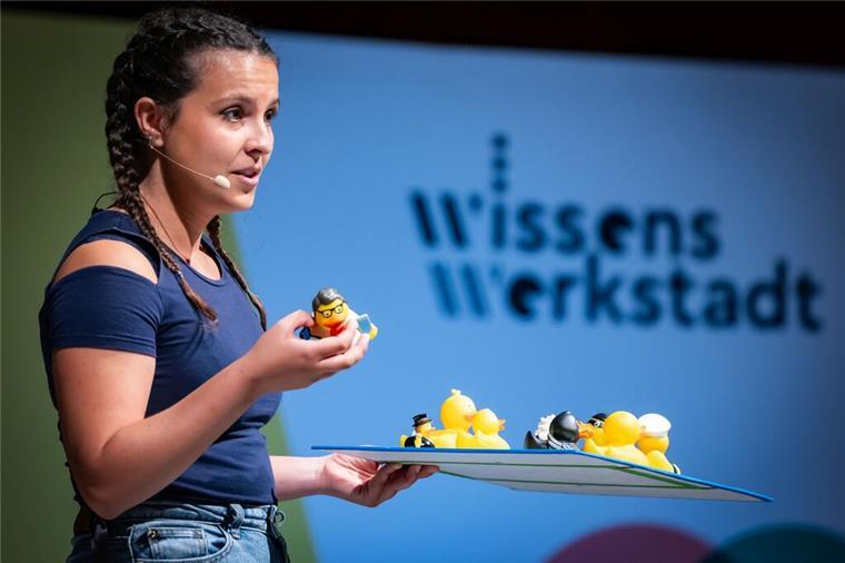 Eine junge Frau hält kleines Gummienten-Spielzeug in der Hand und präsentiert ein Tablett mit mehreren Gummienten-Spielzeugen vor einem Wissens Werkstadt-Schild."