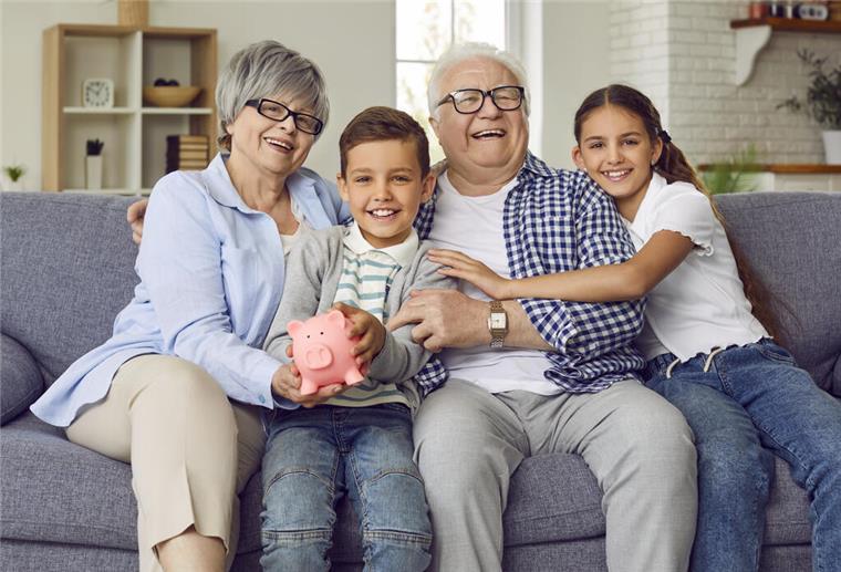 Rentner sitzen mit ihren Enkeln auf dem Sofa und halten ein Sparschwein. Die Bundesregierung plant eine Renten-Reform. Die Frühstart-Rente soll schon 2026 an den Start gehen.