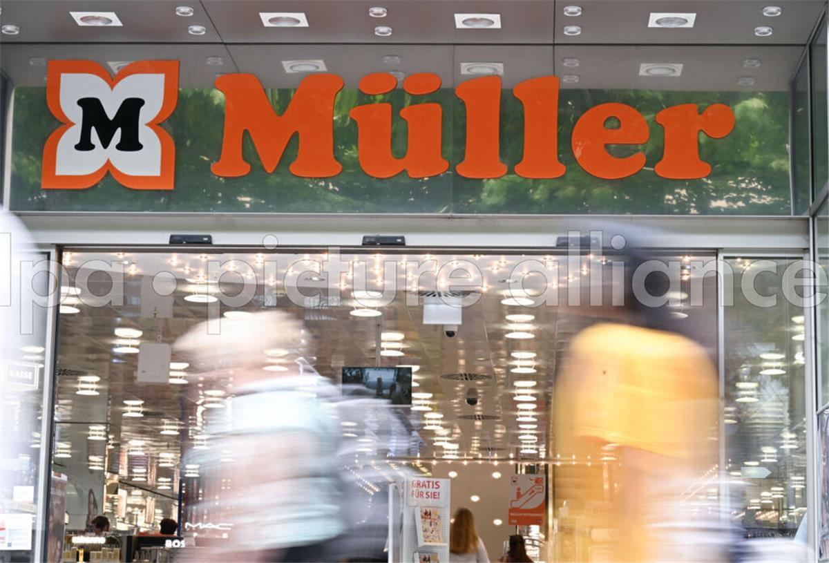 Kunden jubeln! Drogerie-Kette Müller führt neuen Service ein Das Logo der Drogeriemarktkette Müller ist am Eingang einer Filiale angebracht. Im Vordergrund laufen Passanten vorbei