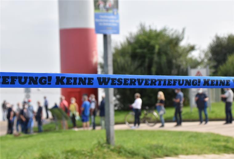 Wesermarsch: Geplante Weservertiefung könnte gestoppt werden Hinter einem blauen Flatterband mit der Aufschrift "Keine Weservertiefung!" sind Menschen und ein Leuchtturm zu sehen.