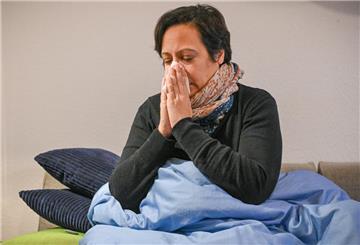 Eine erkältete Frau mit Schnupfen sitzt am 16. Januar 2025 eingewickelt in eine Decke auf einem Sofa und putzt sich die Nase mit einem Taschentuch.