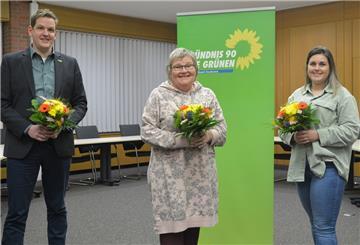 Drei Menschen stehen mit Blumen in der Hand vor einem Banner der Grünen.
