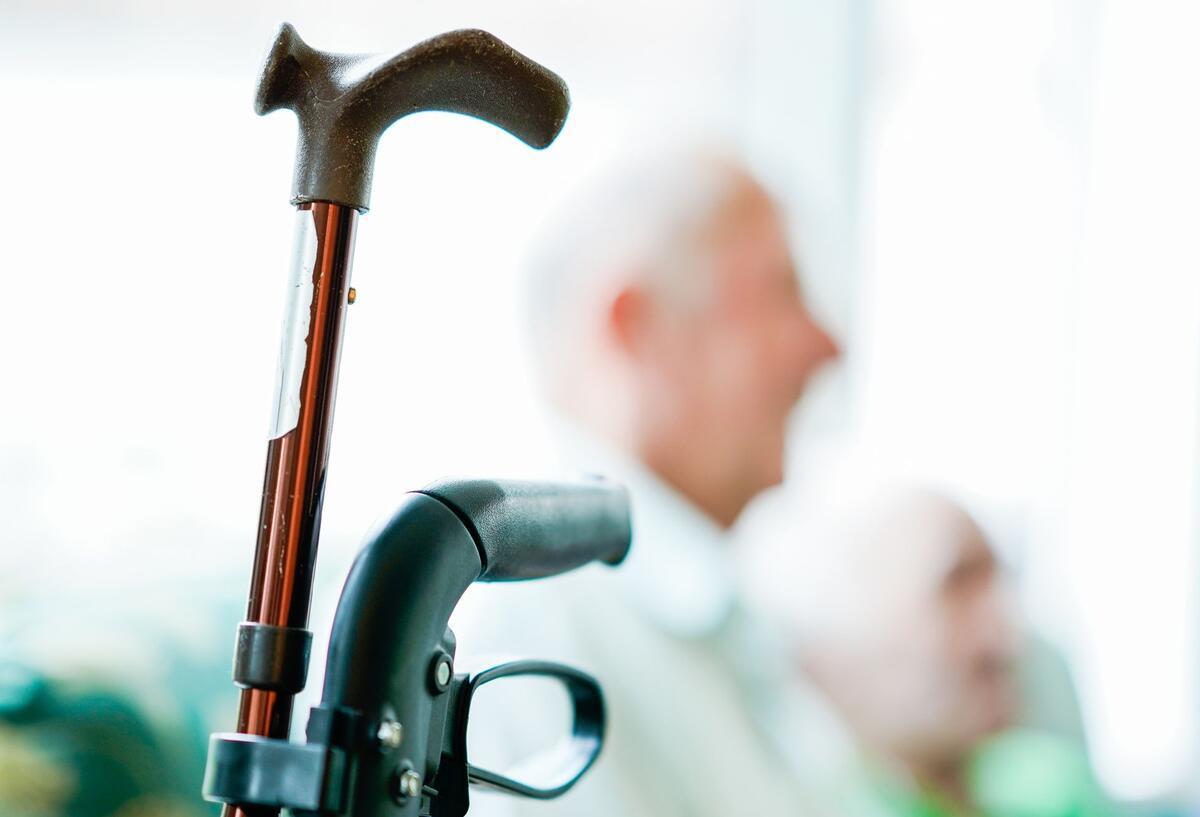 Der Teil eines Rollators und eines Gehstocks. In Hintergrund sieht man unscharf einen Rentner.
