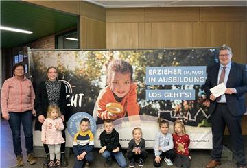 Kinder sitzen vor einem Plakat. Daneben stehen zwei Frauen und ein Mann.