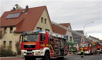 Zwei Feuerwehrwagen stehen in einer Straße. Per Drehleiter versucht ein Feuerwehrmann an einen Schornstein auf einem Haus zu kommen.