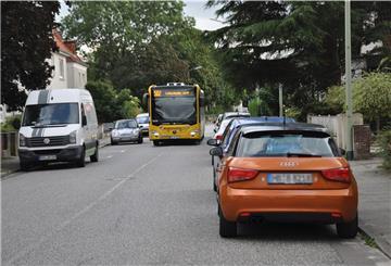 Bus auf einer Straße, dazu abgestellte Autos am Fahrbahnrand.