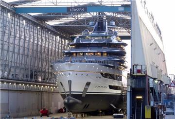 Eine riesige, luxuriöse Megayacht mit glänzendem Rumpf steht im überdachten Schwimmdock der Lürssen-Werft in Bremen-Aumund. Das Schiff ist vollständig gebaut, mehrere Decks mit umlaufenden Balkonen und Antennenaufbauten sind sichtbar. Um das Dock herum stehen technische Anlagen und Gerüste.