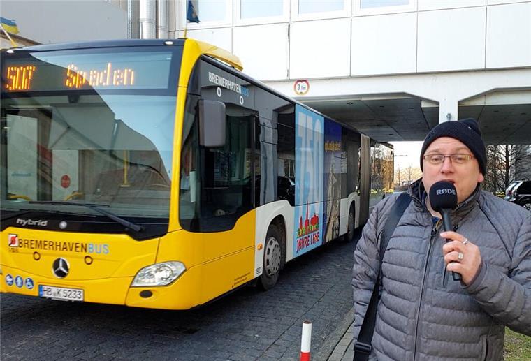 Ein Reporter steht vor einem Bus, der unter einer Unterführung steht. 