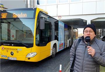 Ein Reporter steht vor einem Bus, der unter einer Unterführung steht. 