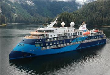 Expeditionskreuzfahrtschiff „Ocean Victory“ fährt durch ruhige Gewässer, umgeben von bewaldeten Bergen.