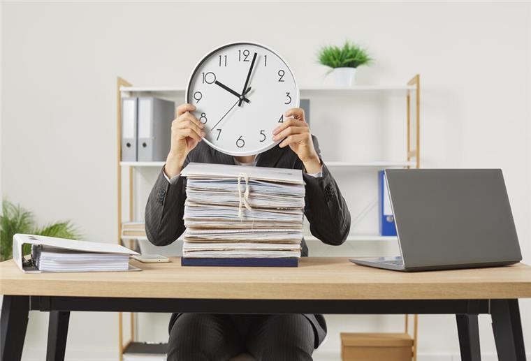 Person in Anzug sitzt an Schreibtisch mit einem großen Stapel Akten und hält eine Uhr vor das Gesicht, die zehn Minuten nach zehn anzeigt; Symbolbild für Zeitdruck oder Überlastung im Büro.