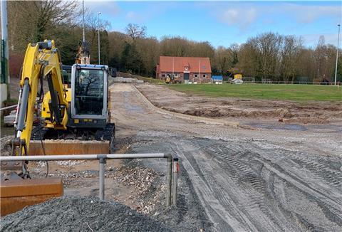 Links steht ein kleiner gelber Bagger, hinter und neben ihm befindet sich eine Sportplatz-Baustelle.