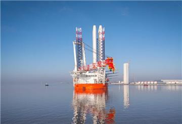 Offshore-Errichterschiff mit leuchtend orangefarbenem Rumpf und großem Kranaufsatz liegt bei klarem Himmel in ruhigem Hafenwasser; im Hintergrund sind Hafenanlagen und Windkraftkomponenten zu sehen.