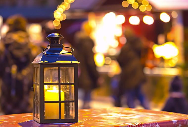 Eine Kerze brennt in einer Laterne. Im Hintergrund sind schemenhaft Buden, Lichter und Menschen auf einem Weihnachtsmarkt zu erkennen.