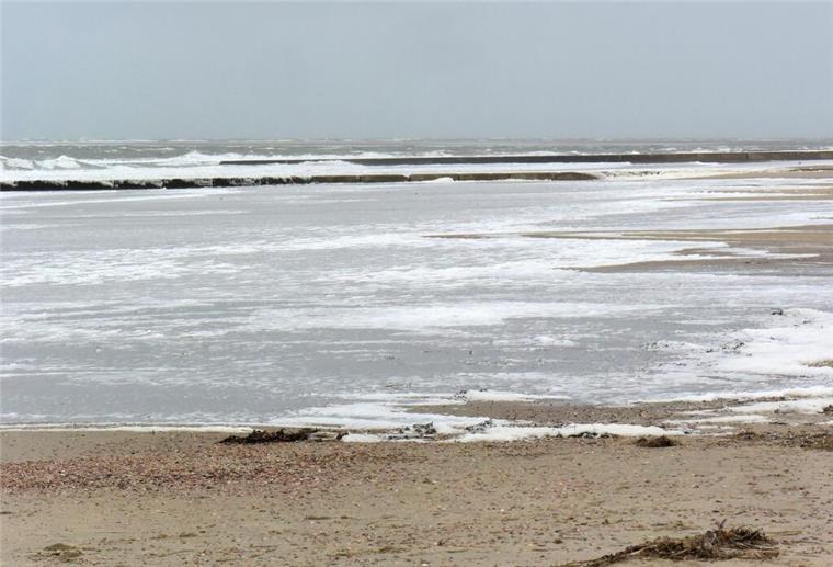 Eine Spaziergängerin geht an der mit Meeresschaum bedeckten Wasserkante am Südbad auf der ostfriesischen Insel.