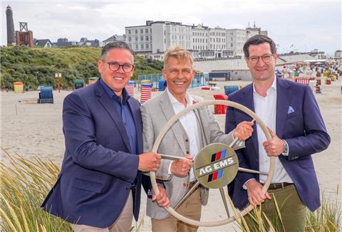 Drei Männer an einem Strand halten ein Steuer in den Händen.  Dr. Bernhard Brons (Mitte) verabschiedet sich nach 38 Jahren bei der AG EMS – das Ruder übernehmen Dr. Wessel Brons (links) und Warner Warnders.