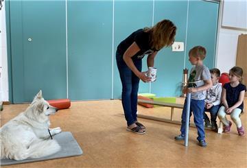 Ein weißer Hund liegt am Boden. Etwas weiter rechts, steht eine Frau mit einer Dose und ein Kind, dass auf die Dose schaut.