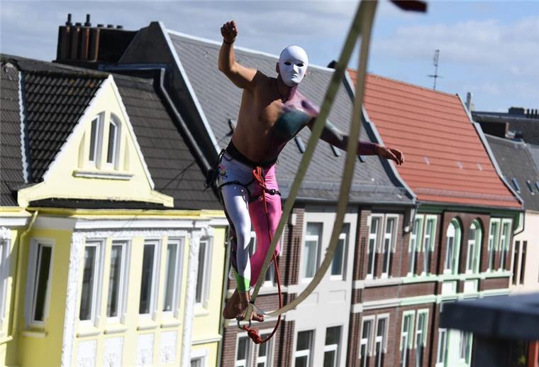 Ein Akrobat jongliert hoch über einer Straße zwischen Altbauten auf einem Seil