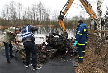 Drei Männer untersuchen die Überreste eines Autos an einem See. Daneben steht ein Bagger.
